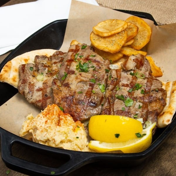 Grilled pork chops with pita, hummus, lemon, and crispy potato slices in a black pan.