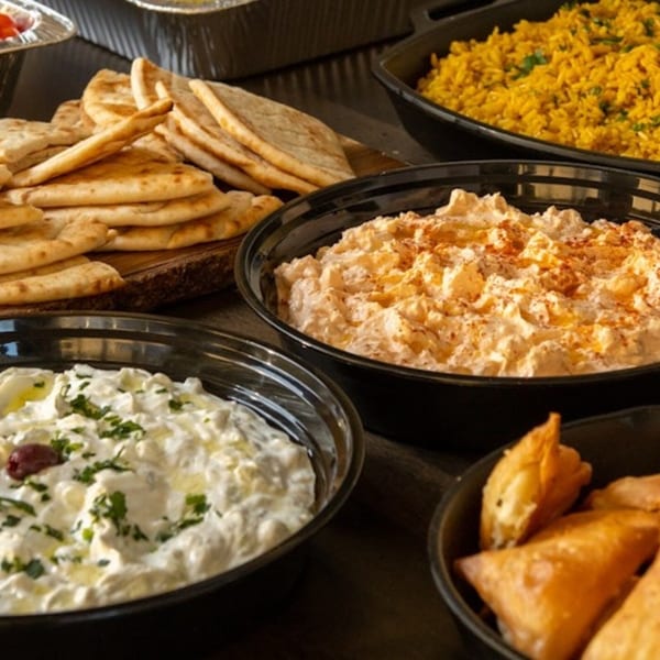 Greek pita bread, tzatziki, hummus, yellow rice, and samosas in black bowls for catering.