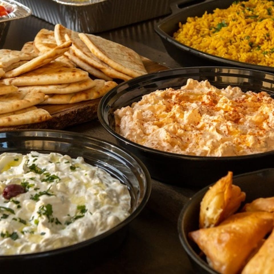 Greek pita bread, tzatziki, hummus, yellow rice, and samosas in black bowls for catering.