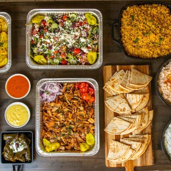 Overhead view of a Souvlaqueria catering spread with Greek salad, chicken, rice, pita, and dips.