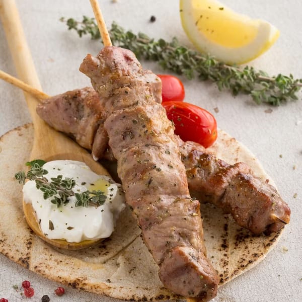 Two Souvlaqueria pork souvlaki skewers on pita bread with tzatziki, cherry tomatoes, lemon, and herbs.