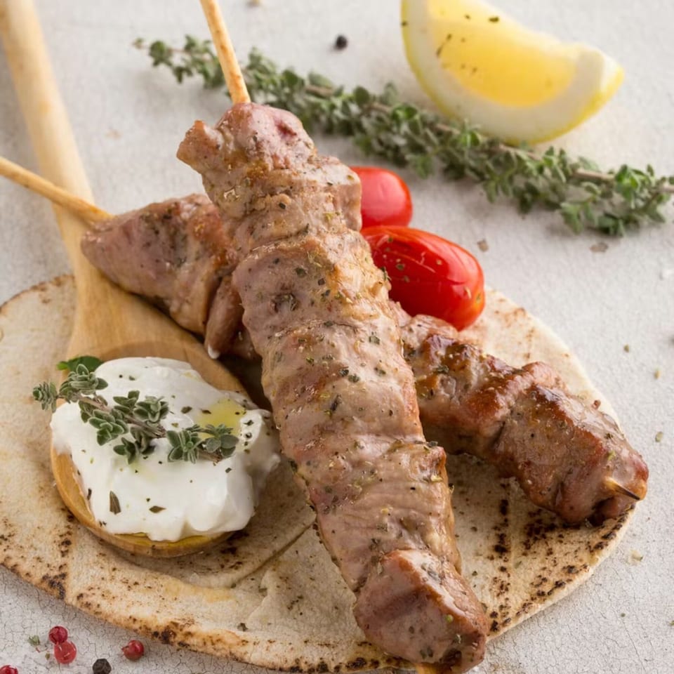 Two Souvlaqueria pork souvlaki skewers on pita bread with tzatziki, cherry tomatoes, lemon, and herbs.