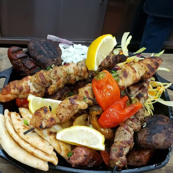 A large platter of grilled Greek meats, including skewers, sausages, and patties, with pita and lemon.