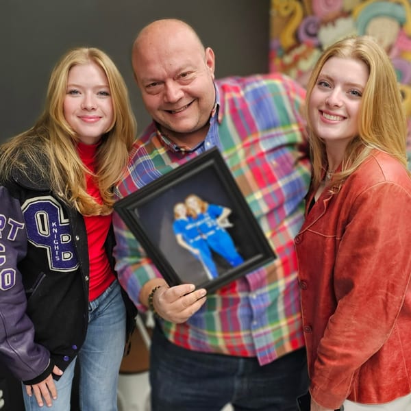A man in a plaid shirt holds a framed photo of two women, flanked by two smiling women.