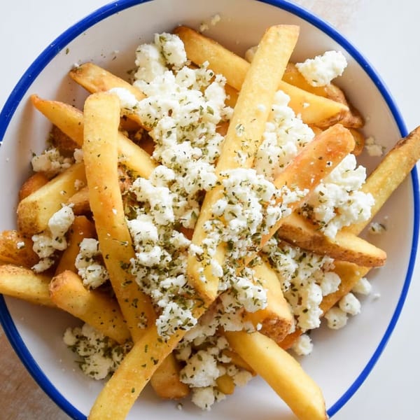 Golden Souvlaqueria fries topped with crumbled feta cheese and dried herbs in a white bowl.