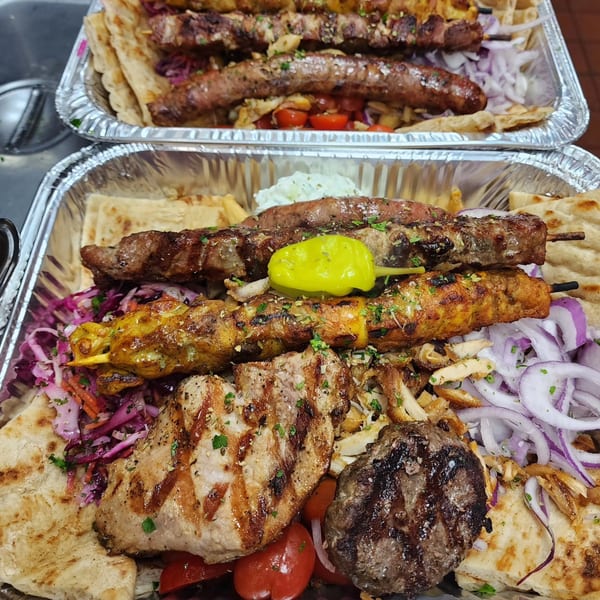 Two foil containers overflowing with grilled Greek meats, pita, red onions, and a yellow pepper.
