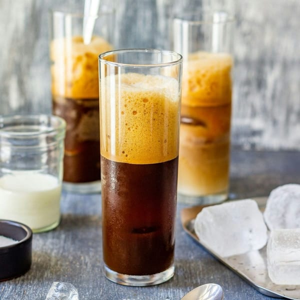 Three tall glasses of Greek frappe coffee with foam, milk, and ice cubes on a table.
