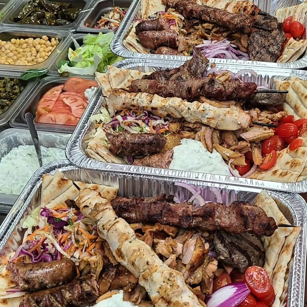 Three aluminum trays filled with Greek Souvlaqueria meats, pita, tzatziki, and various toppings.