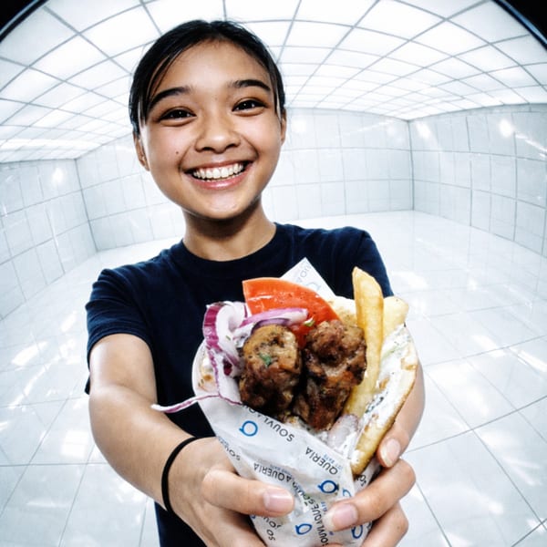 A smiling person holds out a Souvlaqueria gyro with meatballs, fries, and vegetables.