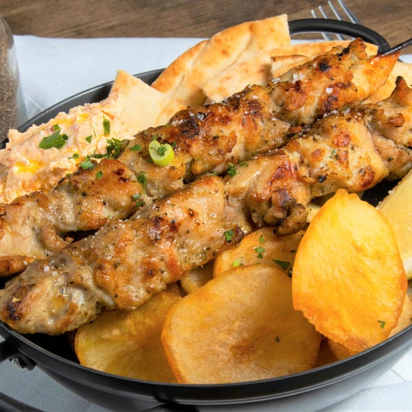 Two chicken souvlaki skewers with pita bread, hummus, and fried potatoes in a black bowl.