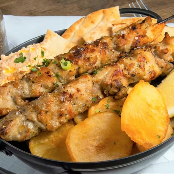 Grilled chicken souvlaki skewers with pita bread, hummus, lemon, and roasted potatoes in a black bowl.