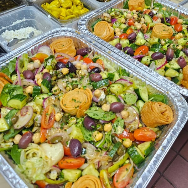 Two foil trays of Souvlaqueria's Greek salad with olives, tomatoes, cucumbers, and pastry swirls.