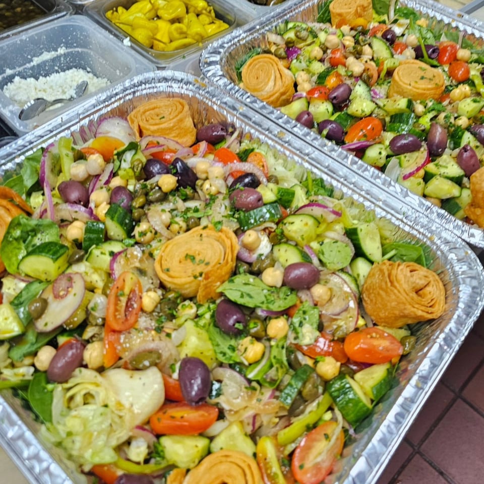 Two foil trays of Souvlaqueria's Greek salad with olives, tomatoes, cucumbers, and pastry swirls.