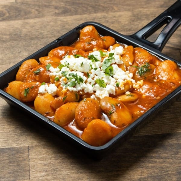 Greek potatoes in tomato sauce, topped with feta cheese and parsley, in a black pan.