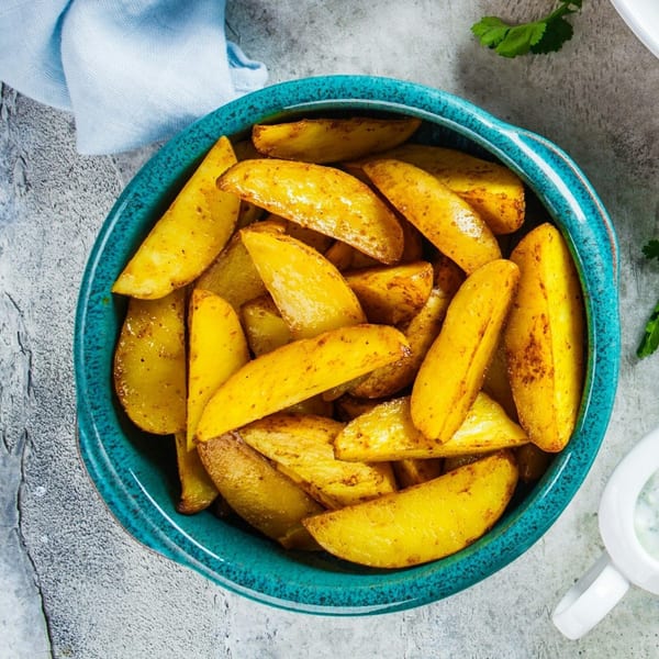 Golden-brown Greek potato wedges seasoned with spices, served in a vibrant teal bowl on a gray surface.