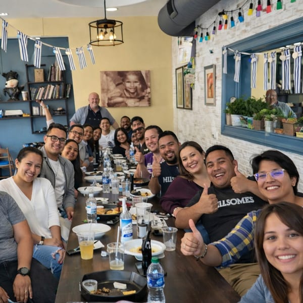 A large group of happy people giving thumbs up at a long table in Souvlaqueria, decorated with Greek flags.