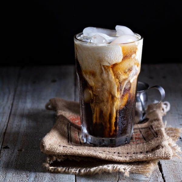 Iced coffee with cream and ice in a tall glass on a burlap coaster and wooden table.